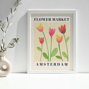 May include: A framed art print featuring a watercolor illustration of tulips in shades of red, pink, and yellow. The words "FLOWER MARKET" and "AMSTERDAM" are printed in navy blue. A beige vase with a unique design holds olive branches.