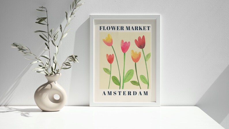 May include: A framed art print featuring a watercolor illustration of tulips in shades of red, pink, and yellow. The words "FLOWER MARKET" and "AMSTERDAM" are printed in navy blue. A beige vase with a unique design holds olive branches.