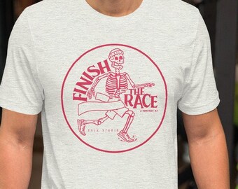 May include: A light gray t-shirt featuring a circular design with the words "Finish the Race" and a skeleton runner. The design is in a reddish-pink color. The shirt is made of a soft material.