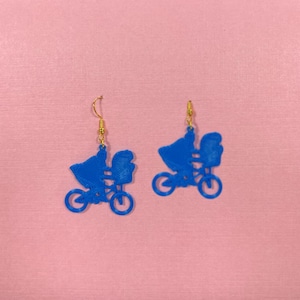 May include: A pair of blue earrings shaped like a silhouette of a person riding a bicycle with a baby in a carrier on the back.