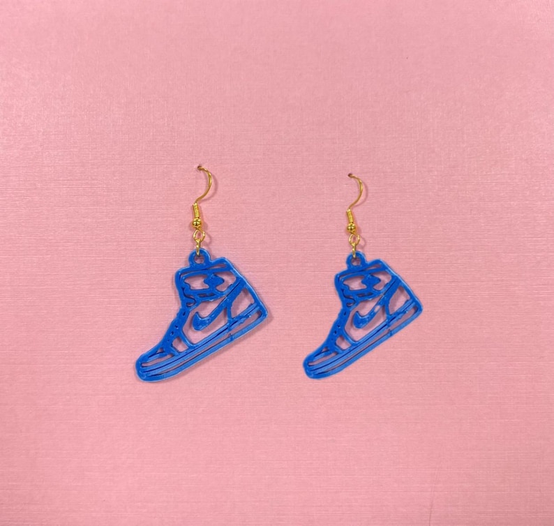 May include: A pair of blue sneaker-shaped earrings with gold-colored ear wires. The earrings are a vibrant blue and feature a cutout design of a high-top sneaker. The background is a solid pink color.