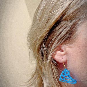 May include: A blue sneaker-shaped earring hangs from a gold-colored hook. The earring is worn by a person with blonde hair. The earring is a cutout design, showing the shape of a high-top sneaker.