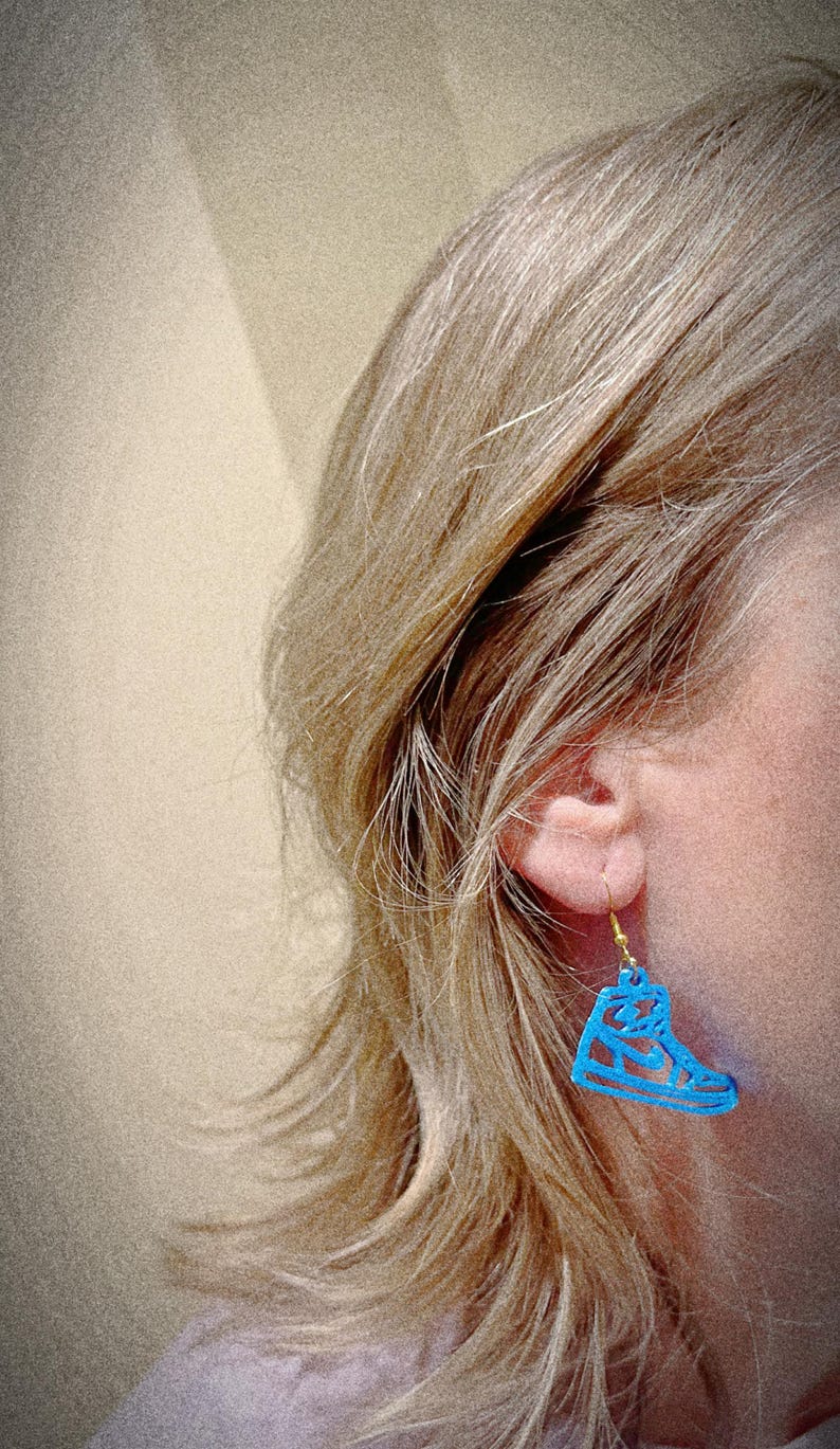 May include: A blue sneaker-shaped earring hangs from a gold-colored hook. The earring is worn by a person with blonde hair. The earring is a cutout design, showing the shape of a high-top sneaker.
