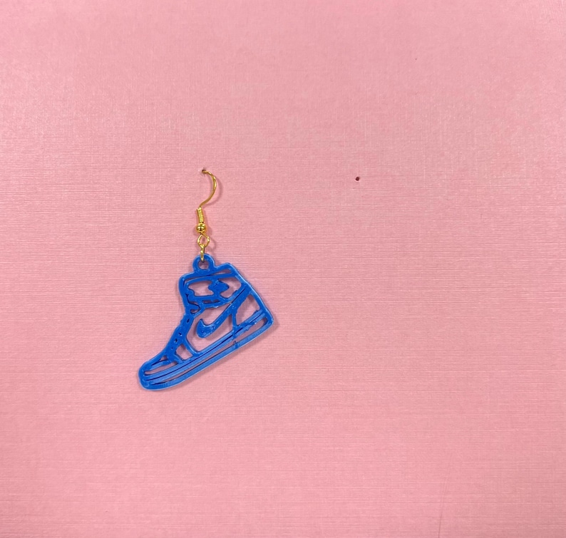 May include: A single blue sneaker-shaped earring with a gold-colored hook. The earring is a cutout design, showing the outline of a high-top sneaker. The background is a solid pink color.