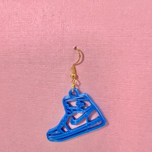 May include: A single blue sneaker-shaped earring with a gold-colored hook. The earring is a cutout design, showing the outline of a high-top sneaker. The earring is hanging against a pink background.
