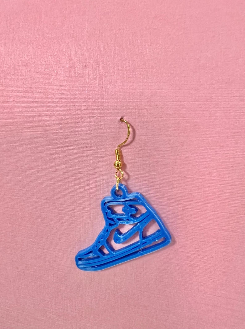 May include: A single blue sneaker-shaped earring with a gold-colored hook. The earring is a cutout design, showing the outline of a high-top sneaker. The earring is hanging against a pink background.
