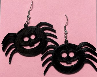May include: A pair of black spider earrings with a smiling face. The earrings are made of plastic and have silver hooks.