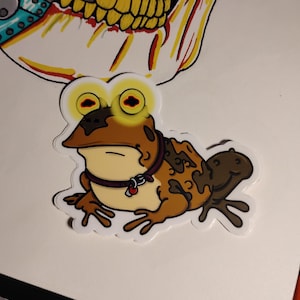 May include: A die-cut sticker of a brown toad with a cream-colored belly, wearing a dark purple collar with a small red tag. The toad has large, glowing yellow eyes with dark pupils. Above the toad is a partial illustration of a yellow skull.