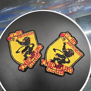 Puede incluir: Dos parches bordados con fondo amarillo y texto negro y rojo. Los parches dicen "Clan Buchanan Throwing Club". Los parches presentan una silueta negra de una persona lanzando una pelota.