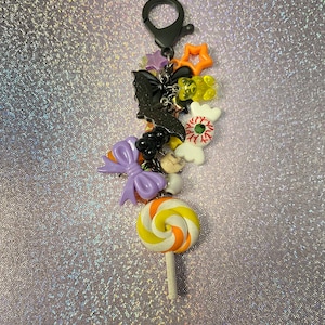 May include: A Halloween-themed keychain with a variety of charms, including a bat, a candy corn lollipop, a ghost, an eyeball, and a star. The charms are attached to a black metal ring with a silver key ring.