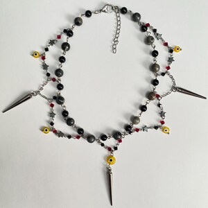 May include: A layered necklace featuring black and gray beads, red accents, and silver star charms. The necklace includes yellow evil eye beads and silver cone pendants. The necklace is on a white background.
