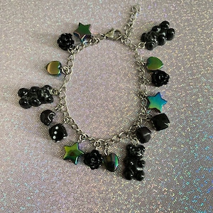May include: A silver charm bracelet featuring various black charms, including a gummy bear, roses, stars, hearts, and dice. Some charms have an iridescent finish. The bracelet is displayed on a sparkly, light purple surface.