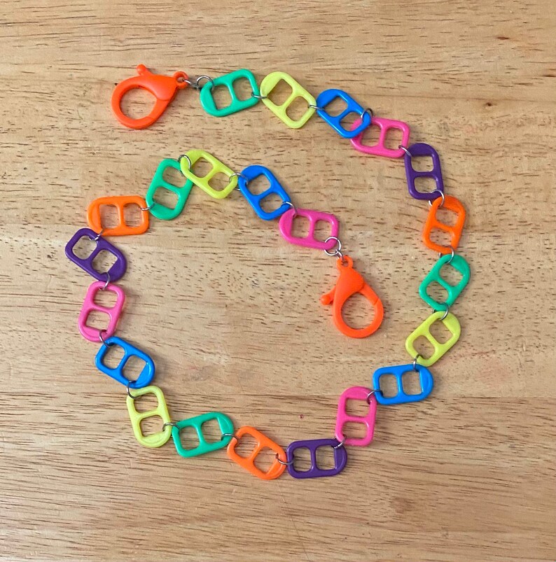 May include: A colorful chain necklace made from linked pull tabs. The chain is made of different colors, including yellow, orange, green, blue, and purple. The chain has two silver clasps.