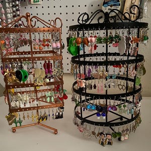 May include: Two earring display stands filled with various earrings. One stand is copper-colored and rectangular, the other is black and circular. The earrings are in many shapes and colors, including fruits, animals, and abstract designs.