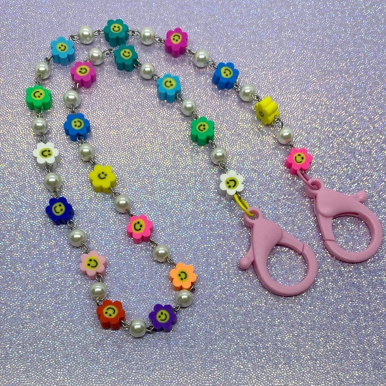 May include: A colorful beaded chain with smiley face charms and pink plastic clasps. The chain is made of white pearls and colorful plastic flowers with yellow smiley faces.