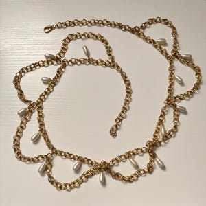 May include: A gold-tone chain belt featuring a series of interconnected circular links arranged in a draped, scalloped pattern. Small, white, teardrop-shaped faux pearls are suspended at regular intervals along the length of the metal chain.