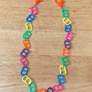 May include: A colorful necklace made from linked pull tabs. The necklace is made of bright colors, including yellow, green, blue, orange, and purple. The necklace has two orange clasps at the ends.
