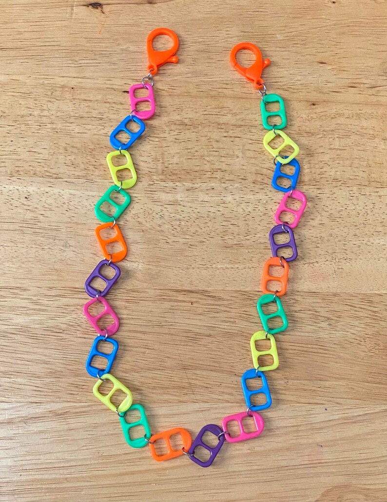 May include: A colorful necklace made from linked pull tabs. The necklace is made of bright colors, including yellow, green, blue, orange, and purple. The necklace has two orange clasps at the ends.