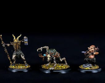 May include: Three miniature fantasy figures painted in a detailed style. The figures are on circular bases and are posed in different actions. The figure on the left is holding a staff and has a horned mask. The figure in the center is a skeletal creature with antlers and is holding a glowing orb. The figure on the right is a small creature with a bow and arrow.