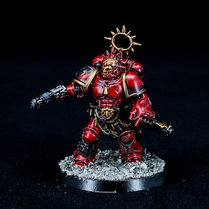 May include: A detailed miniature figure of a warrior in red armor with gold accents. The figure is holding a weapon and stands on a textured base. A golden halo-like structure is above the head. The figure is set against a dark background.