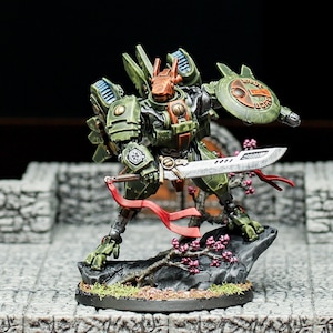 May include: A green and orange painted miniature robot figure holding a silver sword and a green shield with a red and white symbol. The robot is standing on a black base with a small tree and pink flowers.