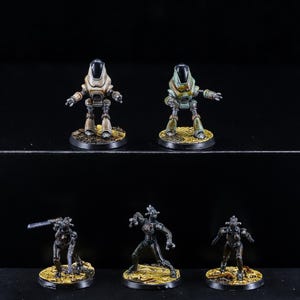 Pode incluir: Miniaturas de robôs em várias poses e cores, incluindo bege, verde e preto. Os robôs estão em bases circulares com uma superfície texturizada de cor dourada. A imagem está em um fundo preto.