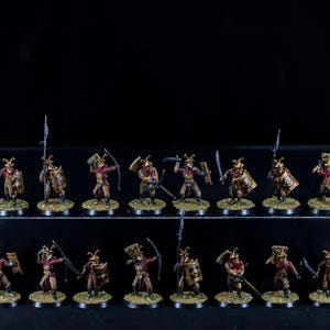 May include: A collection of 18 painted miniature figures, each depicting a warrior with a horned helmet, shield, and weapon. The figures are arranged in three rows on a black surface. The figures are painted in shades of red, gold, and brown.