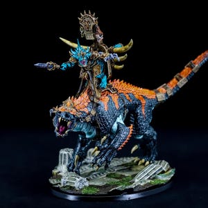 May include: A blue and gold painted miniature of a lizard-like creature with a rider. The rider is a blue and gold painted miniature of a humanoid figure with a sword and a crown. The miniature is set on a base with a rocky terrain.