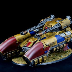 May include: A detailed miniature war tank model, painted in gold, red, and blue. The tank features two large cannons and is mounted on a textured base. The model has intricate details and is set against a black background.