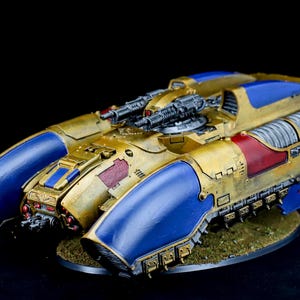 Puede incluir: Modelo en miniatura detallado de un vehículo futurista, principalmente dorado con detalles azules y rojos. El modelo presenta dos cañones prominentes y está montado sobre una pequeña base texturizada. El diseño sugiere un tema de ciencia ficción o fantasía.