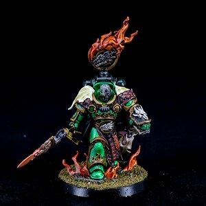 May include: A detailed miniature figure of a warrior in green and gold armor, holding a sword. The figure has a flaming headpiece and is standing on a base with flames and foliage. The figure is set against a black background.