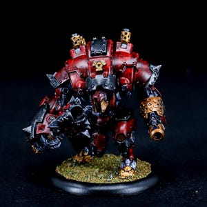 May include: A detailed miniature robot figure, mainly red and black, with gold detailing. The figure stands on a circular base with a textured, grassy surface. The robot has a complex, mechanical design with visible weaponry.