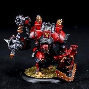 May include: A detailed miniature robot figure, predominantly red and black, with a hammer, shield, and mechanical elements. The figure is posed on a small, textured base, showcasing intricate detailing.