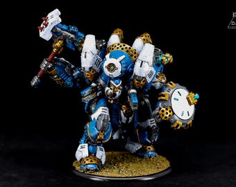 May include: A detailed miniature robot figure, predominantly blue and white, with gold accents. It features a hammer and a shield, standing on a small, textured base. The figure has intricate mechanical details and is set against a black background.
