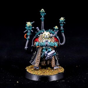 May include: Miniature figurine of a character in a red, black, and gold robe, with teal and silver mechanical attachments. The figure stands on a circular base with a textured surface. The image has a dark background.