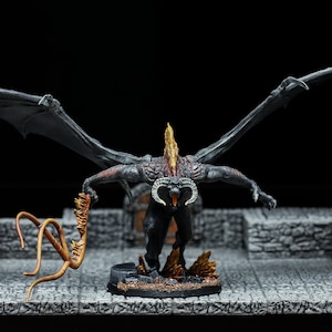 Puede incluir: Una miniatura de dragón negro y rojo con llamas naranjas pintadas en su espalda y alas. El dragón sostiene un látigo dorado en su mano derecha y está de pie sobre una base negra con textura de piedra gris.