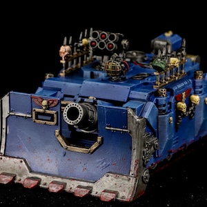 May include: A detailed miniature blue tank model with a large cannon and various weapon attachments. The model features skull and missile details, with a weathered, battle-worn appearance. The model is set against a black background.
