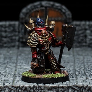 Puede incluir: Una figura en miniatura roja y negra de un marine espacial con un casco adornado con calaveras y un hacha grande. La figura está de pie sobre una pequeña base verde.