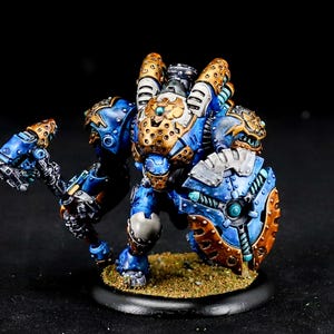 May include: A miniature robot figure with blue, gold, and silver armour. It holds a hammer and shield, standing on a textured base. The figure has a mechanical design, with intricate details and a dark background.