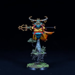 May include: A painted miniature figure of a fantasy creature with horns, a blue and gold robe, and a staff. The figure is standing on a rocky base with green foliage.