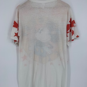 Op de afbeelding: Wit T-shirt met een rood, wit en blauw sterrenpatroon. Het shirt heeft een vervaagde afbeelding van een cartoonfiguur met een blauw en geel ontwerp.