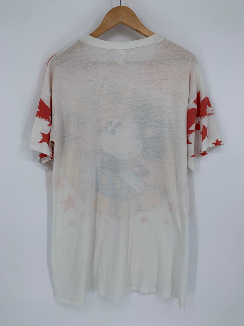 Op de afbeelding: Wit T-shirt met een rood, wit en blauw sterrenpatroon. Het shirt heeft een vervaagde afbeelding van een cartoonfiguur met een blauw en geel ontwerp.