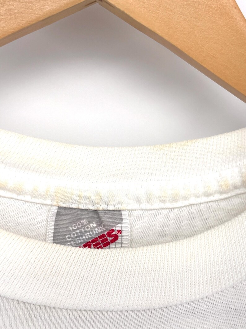 May include: A white t-shirt with a ribbed crew neckline showing signs of light yellow discoloration. A grey fabric tag is sewn into the inner collar, featuring the red text Hanes and the white text 100% Cotton Preshrunk on a grid background.