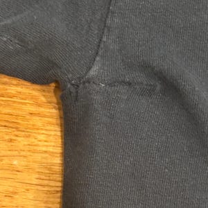 May include: A close-up view of the underarm area of a black t-shirt, showing the seam where the sleeve attaches to the main body of the garment. The fabric appears to be a standard cotton knit with visible stitching along the armhole seam.