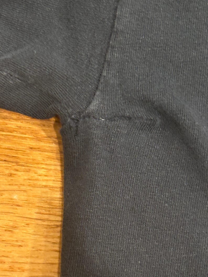 May include: A close-up view of the underarm area of a black t-shirt, showing the seam where the sleeve attaches to the main body of the garment. The fabric appears to be a standard cotton knit with visible stitching along the armhole seam.