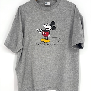 May include: A heather gray short-sleeve t-shirt featuring a graphic of Mickey Mouse pointing to the side with the text "I'M WITH GOOFY" printed underneath in black. The shirt has a crew neckline and is displayed on a wooden hanger.