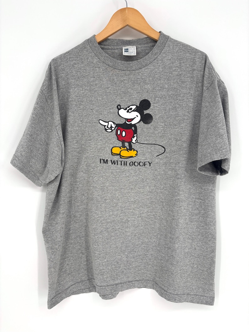 May include: A heather gray short-sleeve t-shirt featuring a graphic of Mickey Mouse pointing to the side with the text "I'M WITH GOOFY" printed underneath in black. The shirt has a crew neckline and is displayed on a wooden hanger.