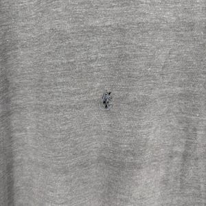 K&ouml;nnte beinhalten: Ein Kleidungsst&uuml;ck aus meliertem grauem Stoff mit einem kleinen, dunklen, ausgefransten Loch in der Mitte des Materials. Die Textur des Stoffes ist sichtbar und zeigt eine Mischung aus hellen und dunklen grauen Fasern auf der Textiloberfl&auml;che.