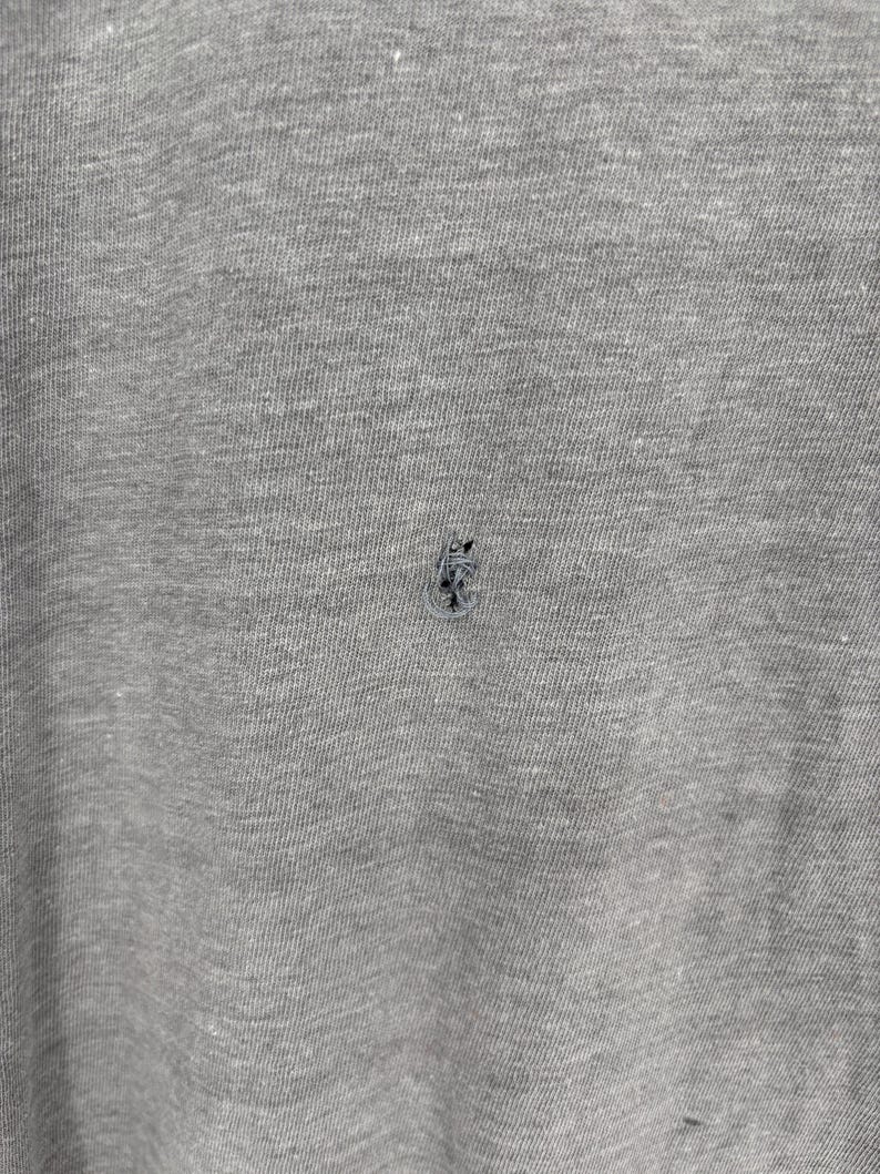 K&ouml;nnte beinhalten: Ein Kleidungsst&uuml;ck aus meliertem grauem Stoff mit einem kleinen, dunklen, ausgefransten Loch in der Mitte des Materials. Die Textur des Stoffes ist sichtbar und zeigt eine Mischung aus hellen und dunklen grauen Fasern auf der Textiloberfl&auml;che.