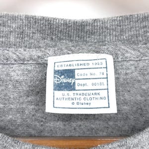 May include: A heather gray t-shirt featuring a white rectangular interior neck label with blue text that reads Established 1923, Disney, Code No. 78, Dept. 0018L, U.S. Trademark Authentic Clothing, and the Disney logo with a copyright symbol.